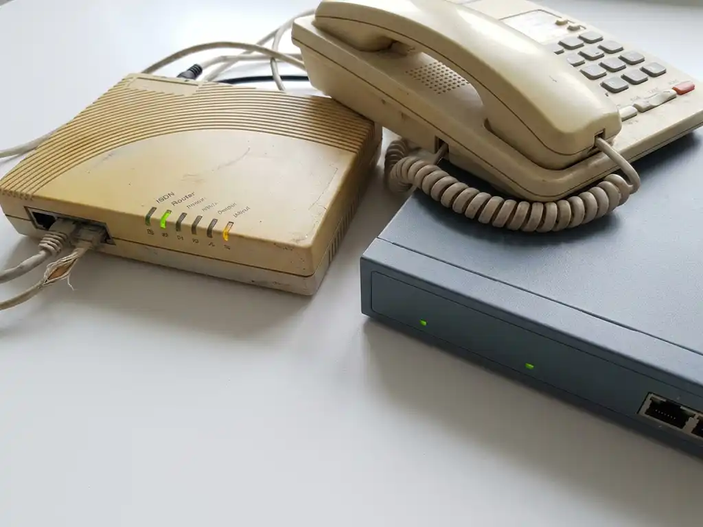 Verouderde ISDN-router en kantoortelefoon uit de jaren 90 naast een moderne netwerkswitch op wit oppervlak.